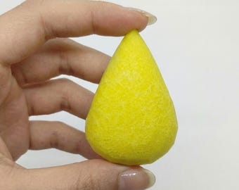 May include: A bright yellow, teardrop-shaped makeup sponge. The sponge has a textured surface and is held between fingers against a white background. The sponge is a vibrant, solid color.