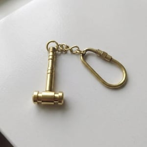 Pode incluir: Um pequeno porta-chaves em forma de martelo dourado. A cabeça e o cabo do martelo são cilíndricos, com uma pequena corrente e um mosquetão. O porta-chaves é feito de metal e tem um acabamento brilhante. Mede cerca de 4 cm de comprimento.