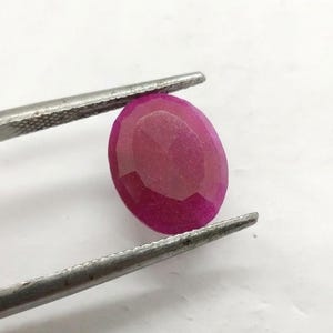 Könnte beinhalten: Ein facettierter, ovaler Rubin-Edelstein, der von einer silbernen Pinzette gehalten wird. Der Rubin hat eine tiefe, satte rosa Farbe, mit mehreren Facetten, die das Licht einfangen. Die Pinzette ist scharf fokussiert.