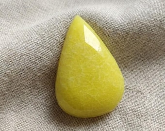May include: A teardrop-shaped, polished gemstone in a vibrant yellow hue. The stone has a smooth, glossy surface with subtle internal veining. The gemstone is set against a neutral, textured fabric background.