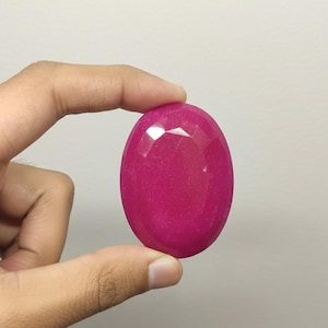 May include: A faceted, oval-shaped, fuchsia gemstone. The polished stone has a smooth surface with multiple facets that catch the light. The gemstone is held between two fingers against a neutral background.