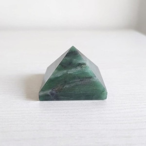 May include: A green pyramid-shaped stone with dark green and white marbling. The pyramid is sitting on a white surface. The pyramid is a decorative item.