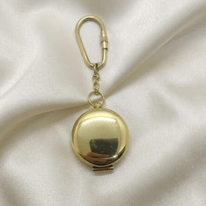 Puede incluir: Un llavero con medallón redondo dorado con una abertura con bisagras. El medallón está unido a una cadena y a un mosquetón. El fondo es una tela blanca y sedosa.