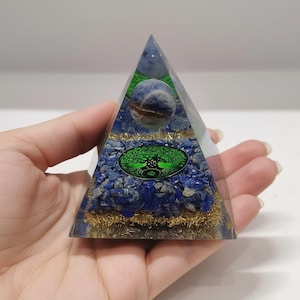 Pode incluir: Uma pirâmide de cristal de orgonite transparente com detalhes azuis e dourados. A pirâmide apresenta um símbolo da árvore da vida e uma esfera azul. A base é preenchida com pedras azuis e metal dourado.