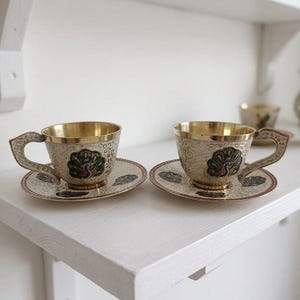 May include: Two ornate teacups and saucers with a peacock design. The cups have a gold interior and handles, with a silver exterior. The saucers have a matching design. The teacups and saucers are displayed on a white shelf.