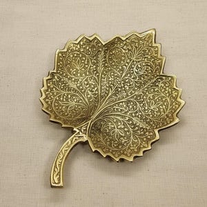 Peut inclure: Un plat décoratif en forme de feuille en laiton avec des motifs floraux et de vigne complexes. Le plat a une tige et une surface texturée. La couleur est dorée et le design évoque la nature, idéal pour la décoration.