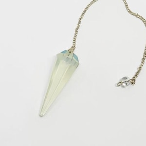 Puede incluir: Un péndulo de cristal claro y facetado con una cadena plateada y una pequeña cuenta transparente en el extremo. El cristal es de color amarillo verdoso pálido y tiene forma puntiaguda en la parte inferior. La cadena mide unos 20 cm.