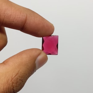May include: A rectangular, faceted, magenta gemstone is held between two fingers against a white background. The gem has a polished surface and appears to be transparent, allowing light to pass through. The gem's shape is a perfect rectangle.