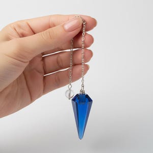 May include: A faceted, deep blue crystal pendulum suspended from a silver chain. A small, clear crystal bead is also attached to the chain. The pendulum is held against a plain white background.