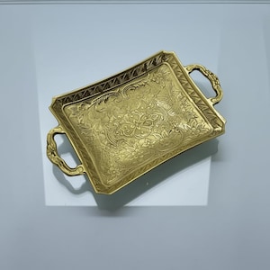 May include: A gold-coloured, rectangular tray with ornate floral and scrollwork detailing. The tray features two curved handles and a decorative border. The surface is highly reflective, and the tray appears to be made of metal.