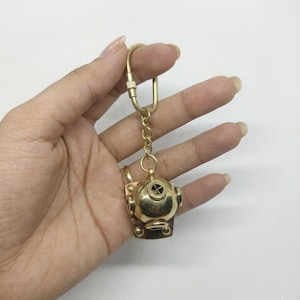 May include: A brass-colored diver's helmet keychain. The miniature helmet has a round top with a cross-shaped detail and several circular side features. It hangs from a gold-colored chain and a large key ring.