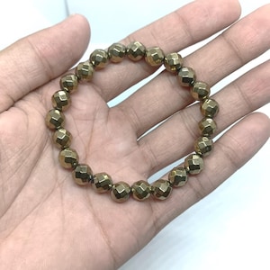 Puede incluir: Una pulsera dorada con cuentas facetadas. Las cuentas están dispuestas en un patrón circular, creando un bucle continuo. La pulsera se sostiene en una mano sobre un fondo blanco.