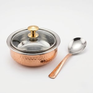 May include: A round, hammered copper serving bowl with a stainless steel interior, featuring a clear glass lid with a gold-toned knob and a matching hammered copper serving spoon with a polished stainless steel bowl.