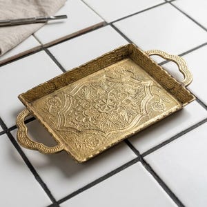 May include: A rectangular gold-colored metal tray featuring an intricate embossed floral pattern across its surface. The tray includes two decorative handles on opposite ends and has a raised, scalloped edge border with a textured finish.