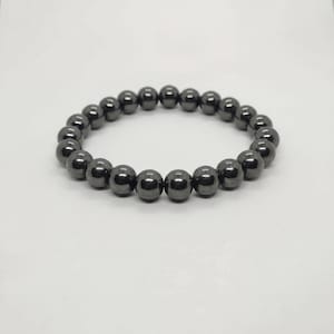 May include: A dark gray beaded bracelet. The bracelet is made of round, polished beads strung together to form a continuous loop. The beads have a metallic sheen and reflect light, creating a subtle shimmer. The bracelet is displayed on a white background.