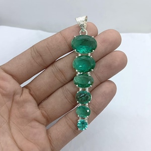 Könnte beinhalten: Ein langer, vertikaler Anhänger mit einer Reihe ovaler und quadratisch geschliffener grüner Edelsteine. Die Edelsteine sind in einem silberfarbenen Metall gefasst, mit einer strukturierten Schlaufe oben für eine Kette. Der Anhänger wird in einer Hand vor weißem Hintergrund gehalten.