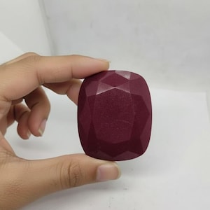 May include: A maroon-coloured, faceted object with a geometric design. The object is held in a hand against a white background. The object's shape is rectangular with rounded corners.