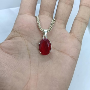 May include: Silver necklace featuring a large, oval-cut, deep red gemstone pendant. The pendant is set in a silver bezel and hangs from a silver chain. The gemstone has a faceted surface, creating a sparkling effect.