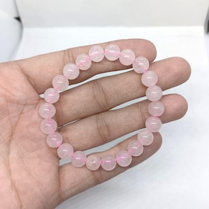 May include: A rose quartz beaded bracelet. The bracelet features round, light pink beads strung together on a pink elastic cord. The beads have a translucent quality, and the bracelet is displayed on a light background.