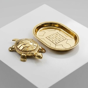 May include: A gold-colored metal turtle figurine and a matching oval-shaped tray. The tray features a beaded rim and an engraved turtle outline containing a three-by-three grid of numbers, specifically 8, 1, 6, 3, 5, 7, 4, 9, and 2.