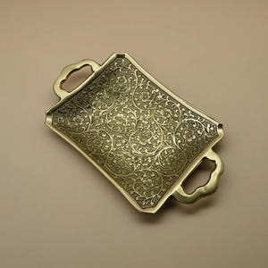 Puede incluir: Una bandeja rectangular de latón con intrincados patrones florales y dos asas curvas. La bandeja tiene una forma ligeramente curvada y un diseño detallado en relieve. El color general es un dorado antiguo cálido.