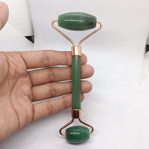 Op de afbeelding: Een groene jade gezichtsroller met een roségoudkleurig metalen frame. De roller heeft twee afgeronde jadestenen, één aan elk uiteinde, verbonden door het metalen frame en een jade handvat. De roller wordt in een hand gehouden.