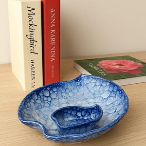 Op de afbeelding: Een blauw en wit keramisch serveerschaaltje met een bubbelpatroon, met een kleiner schaaltje met hetzelfde ontwerp. Twee boeken, "Mockingbird" en "Anna Karenina", en een boek met de titel "Flowers" staan op de achtergrond.