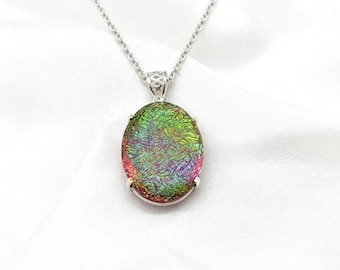 May include: Oval pendant necklace with a dichroic glass pendant in shades of green, red, and purple. The pendant is set in a silver-colored metal frame and hangs from a delicate silver chain. The necklace is displayed on a white background.