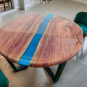May include: A round wooden table with a glossy finish and a vibrant blue resin river running through the center. The table has a dark green metal base and is paired with a teal chair. The table's diameter is approximately 122 cm.