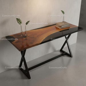 Puede incluir: Una consola moderna con una tapa de madera de borde vivo y una incrustación de resina negra. La mesa tiene una base de metal negro con un diseño en forma de X. Dos jarrones de cristal con hojas verdes están sobre la mesa.