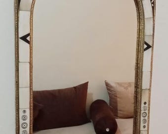 May include: Arched mirror with a decorative frame. The frame features a gold-colored trim, white and black geometric patterns, and a repeating circular design. The mirror reflects a bed with brown pillows.
