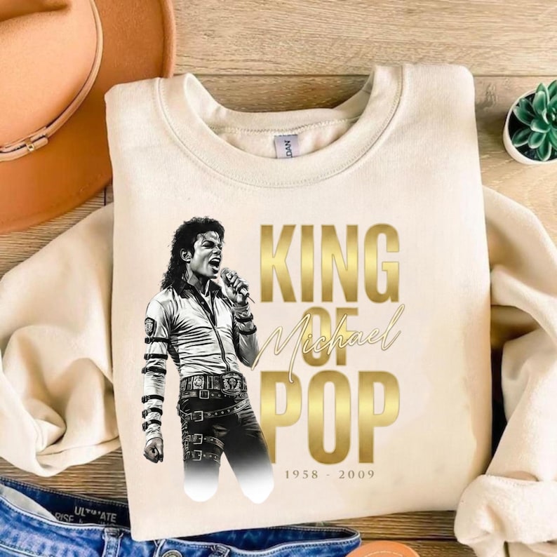 May include: Cream-colored sweatshirt featuring a black and white image of Michael Jackson singing. Gold text reads "KING OF POP" with "Michael" in cursive. The dates "1958 - 2009" are below the text. The sweatshirt is a crew neck style.
