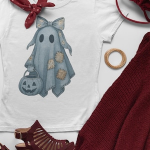 Könnte beinhalten: Weißes T-Shirt mit einer Aquarellillustration eines Geistes in einem blauen Laken, mit Schleife, Patches und einem Kürbis-Eimer. Das Bild enthält einen roten Schal, bordeauxfarbene Absätze und eine passende Handtasche, was auf ein Halloween-Thema hindeutet.