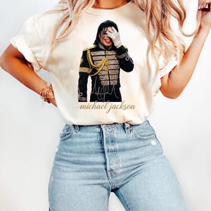 May include: Cream-colored t-shirt featuring a graphic of Michael Jackson in a black military-style jacket with gold accents. The image includes his signature and the text "michael jackson" in a cursive font. The shirt is paired with blue jeans.