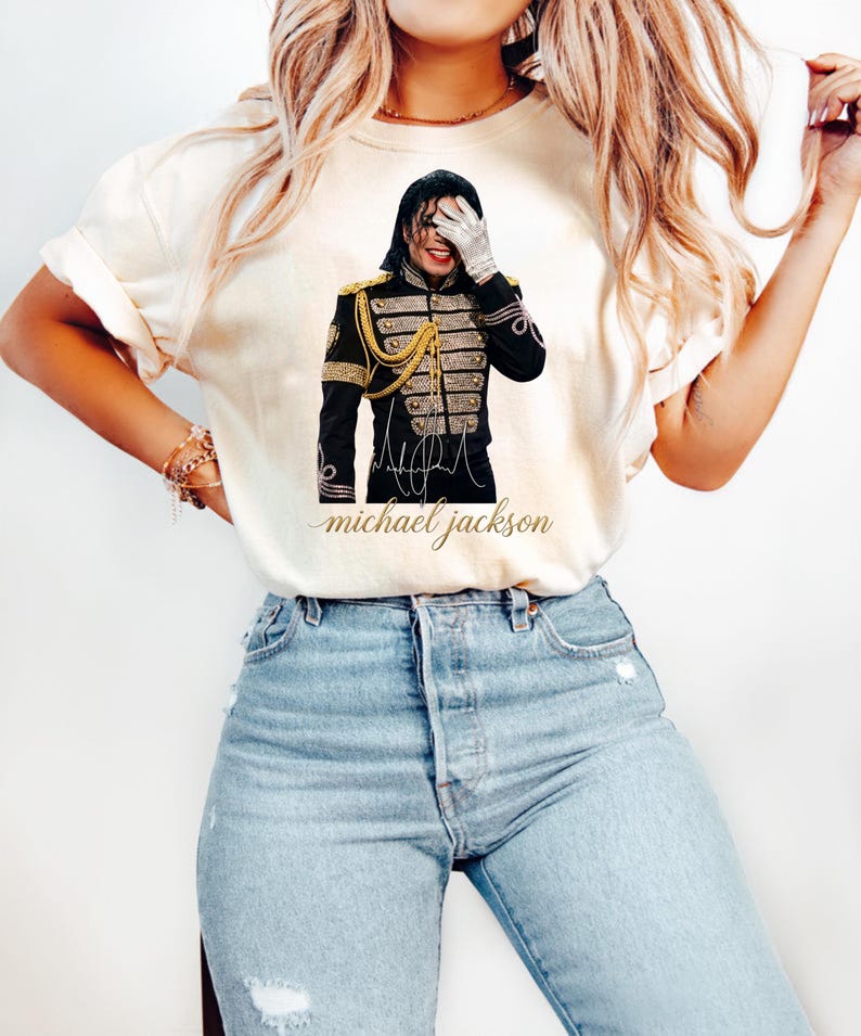 May include: Cream-colored t-shirt featuring a graphic of Michael Jackson in a black military-style jacket with gold accents. The image includes his signature and the text "michael jackson" in a cursive font. The shirt is paired with blue jeans.