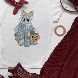 Könnte beinhalten: Weißes T-Shirt mit blauem Geisterdesign, Schleife und Kürbis-Eimer. Der Geist ist mit floralen Mustern verziert. Accessoires: bordeauxfarbener Schal, Schuhe und Handtasche. Halloween-Thema.
