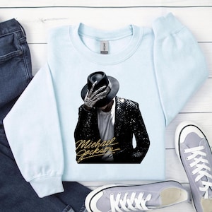 May include: Light blue sweatshirt featuring a graphic of Michael Jackson in a black hat and sequined jacket, with a skeleton hand. The words "Michael Jackson" are written in gold script below the image. The sweatshirt is paired with blue jeans and gray sneakers.