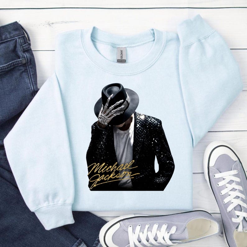 May include: Light blue sweatshirt featuring a graphic of Michael Jackson in a black hat and sequined jacket, with a skeleton hand. The words "Michael Jackson" are written in gold script below the image. The sweatshirt is paired with blue jeans and gray sneakers.