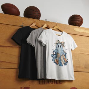 Könnte beinhalten: Drei T-Shirts hängen an Holzbügeln. Ein schwarzes, ein graues und ein weißes T-Shirt. Das weiße T-Shirt hat eine Aquarellillustration eines Geistes, der einen Kürbis-Eimer hält, mit Fledermäusen. Der Geist trägt einen Patchwork-Quilt.