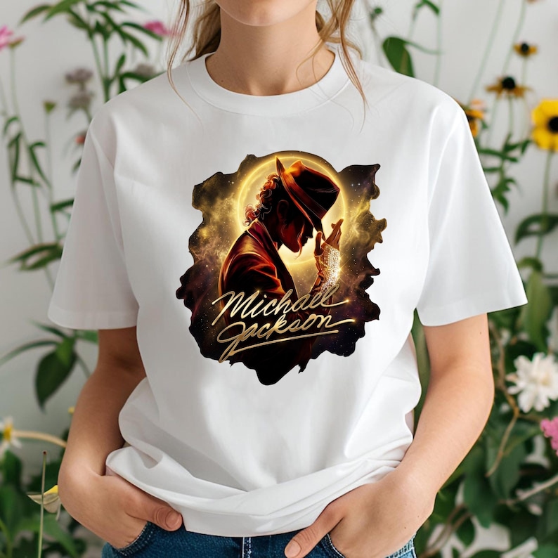 May include: White t-shirt featuring a graphic of Michael Jackson in a hat and suit, set against a golden, cosmic background. The name "Michael Jackson" is written in gold script below the image. The shirt is a classic crew neck.