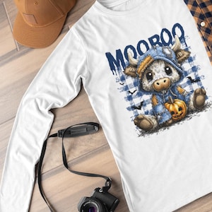 Könnte beinhalten: Weißes Langarmshirt mit einer Cartoon-Kuh, die einen blauen Kapuzenpullover trägt und einen Kürbis hält. Das Shirt trägt den blauen Schriftzug "MOOROO". Eine braune Baseballkappe, eine Kamera und eine Brille sind ebenfalls im Bild.