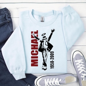 Peut inclure: Sweat-shirt bleu clair avec un motif noir et rouge d'une silhouette dansante, le nom "MICHAEL" en rouge et les ann&eacute;es "1958-2009". Le sweat-shirt est pr&eacute;sent&eacute; avec un jean bleu et des baskets grises.
