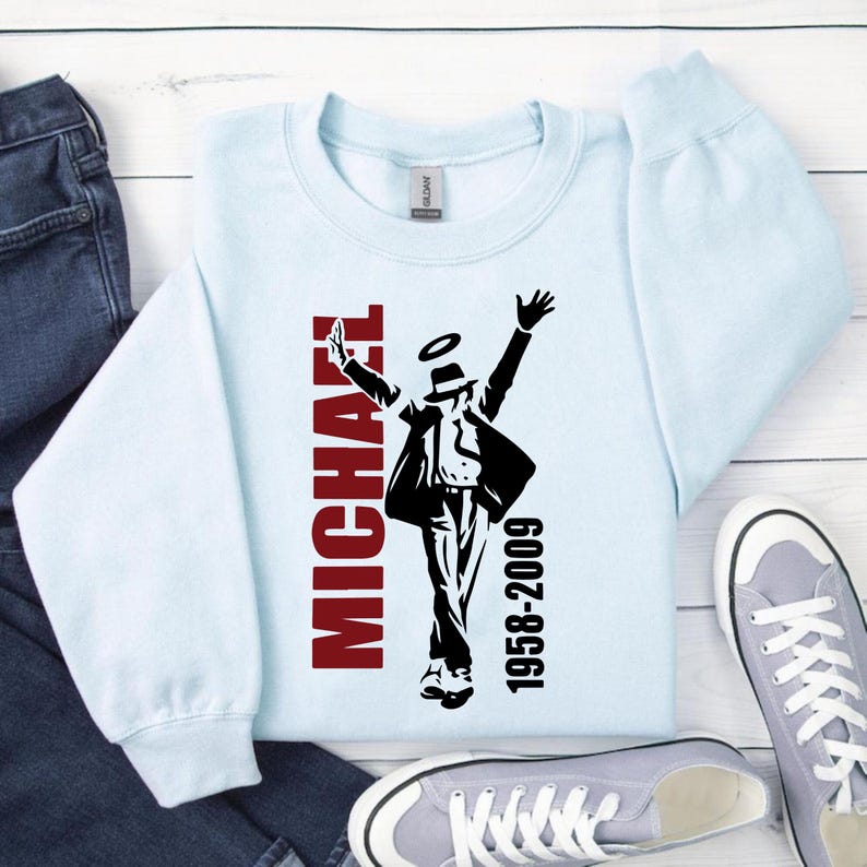 Peut inclure: Sweat-shirt bleu clair avec un motif noir et rouge d'une silhouette dansante, le nom "MICHAEL" en rouge et les ann&eacute;es "1958-2009". Le sweat-shirt est pr&eacute;sent&eacute; avec un jean bleu et des baskets grises.