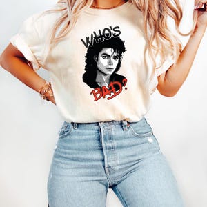 Peut inclure: T-shirt cr&egrave;me avec un graphisme noir et blanc de Michael Jackson et le texte "WHO'S BAD?" en noir et rouge. Le t-shirt est associ&eacute; &agrave; un jean bleu clair.