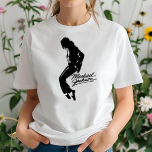 Peut inclure: T-shirt blanc avec une silhouette noire de Michael Jackson dans une pose de danse. Le texte en cursive "Michael Jackson" est &eacute;crit en dessous. Le t-shirt est en mati&egrave;re douce et a un col rond.