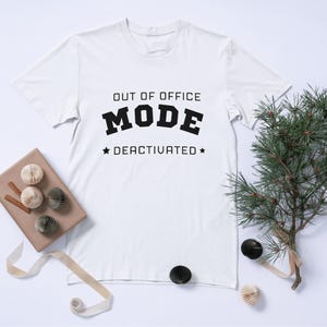 May include: A white short-sleeved t-shirt featuring the black text "OUT OF OFFICE MODE DEACTIVATED" printed on the chest. The design includes the word "MODE" in a bold, blocky font, with a small star symbol on each side of the word "DEACTIVATED".