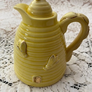 May include: A yellow ceramic creamer with a lid and handle. The creamer has a beehive design with embossed bee accents and gold trim. The spout is curved, and the handle is adorned with gold leaf detailing. The creamer is approximately 6 inches tall.