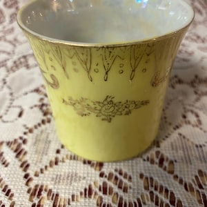 Puede incluir: Una pequeña taza de té amarilla con ribetes dorados y diseños florales. La taza tiene una forma ligeramente acampanada y un interior nacarado. Delicados patrones dorados adornan el exterior, añadiendo un toque de elegancia a esta taza de té vintage.