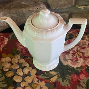 May include: A white porcelain teapot with an octagonal body, a matching lid with a central knob, a curved spout, and a geometric handle. The base and the rim of the lid feature a subtle embossed floral pattern, and the teapot has a glossy finish.