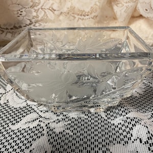 Peut inclure: Un bol carré en verre transparent avec des motifs floraux gravés. Le bol a un bord à facettes et repose sur une surface à motifs blancs et noirs. Le bol est probablement destiné au service ou à l'exposition.